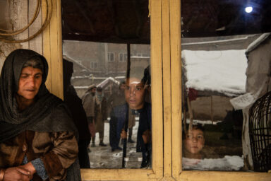 KABUL | KABUL | AFGHANISTAN | 2/6/22 | Latifa (55) stands outside of her home, clay wall and plastic as the roof, in Qala-i Wahed neighborhood of Kabul while her grandchildren look out through the window. 
The family were given refuge on a land after they were displaced from their home province of Parwan and have built a make-shift home from mud, plastic and fabric. The roof of the house collapsed last year after a heavy snowfall.